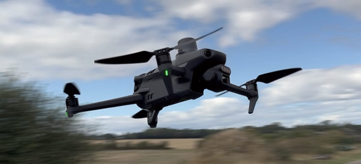 A quadcopter survey drone in flight low over a farm track, a camera payload mounted beneath it, hedgerow and stubble fields blurred behind.